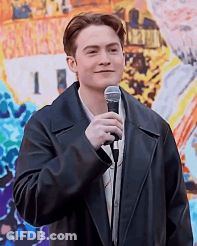 A Young Man In A Black Leather Jacket Is Holding A Microphone In Front Of A Painting . GIF