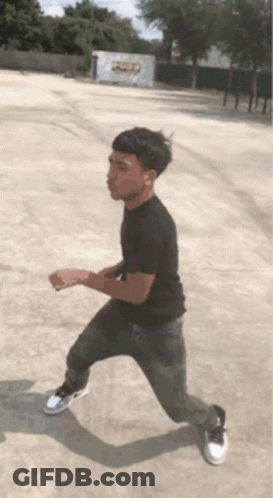 A Young Man In A Black Shirt And Grey Jeans Is Standing In A Parking Lot GIF