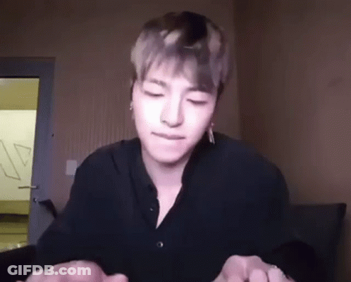 A Young Man In A Black Shirt Is Sitting In A Room With His Eyes Closed And Holding His Hands Together . GIF
