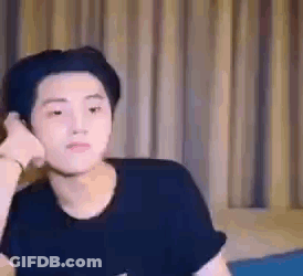 A Young Man In A Black Shirt Is Sitting In Front Of A Curtain And Making A Funny Face . GIF
