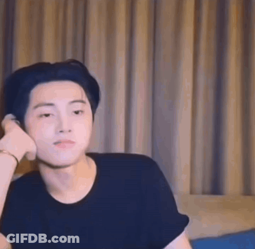 A Young Man In A Black Shirt Is Sitting On A Bed With His Hand On His Ear . GIF