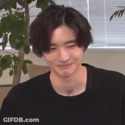 A Young Man In A Black Shirt Is Smiling And Looking At The Camera . GIF