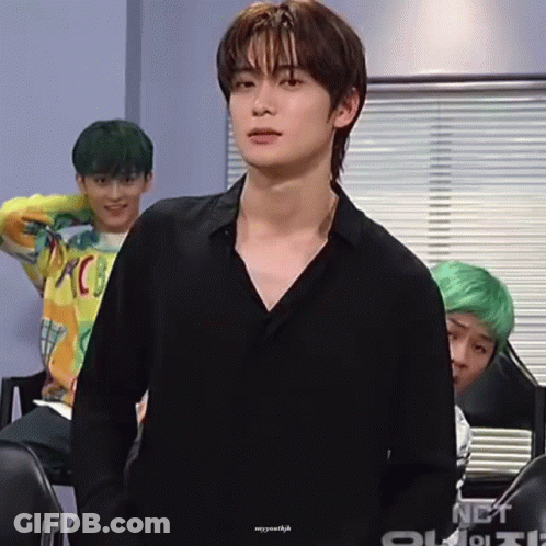 A Young Man In A Black Shirt Is Standing In Front Of A Window With A Group Of Young Men Behind Him . GIF