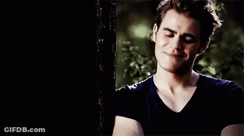 A Young Man In A Black Shirt Is Standing Next To A Tree And Smiling . GIF