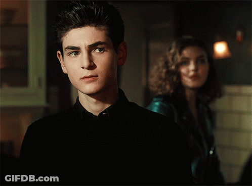 A Young Man In A Black Shirt Stands In Front Of A Woman In A Blue Jacket GIF