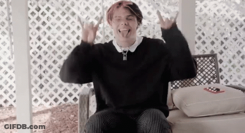 A Young Man In A Black Sweater Is Sitting In A Chair And Making A Devil Horns Sign . GIF