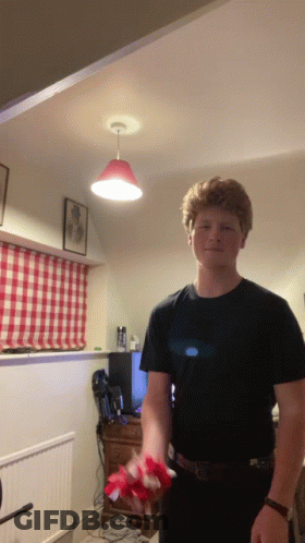 A Young Man In A Black T-Shirt Is Holding Flowers In A Living Room GIF