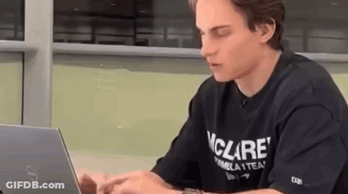 A Young Man In A Black T-Shirt Is Sitting At A Table Using A Laptop Computer . GIF