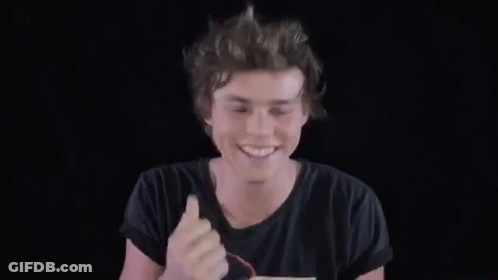 A Young Man In A Black T-Shirt Is Smiling And Waving His Hand In The Air . GIF