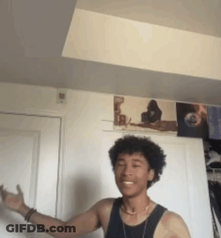 A Young Man In A Black Tank Top Is Standing In A Room With A Picture On The Wall Behind Him . GIF