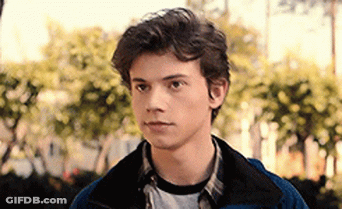 A Young Man In A Blue Jacket And Plaid Shirt Is Standing In Front Of Trees . GIF