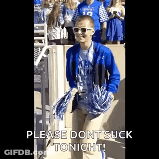 A Young Man In A Blue Jacket And Sunglasses Is Holding Pom Poms And Walking In A Crowd Of People . GIF