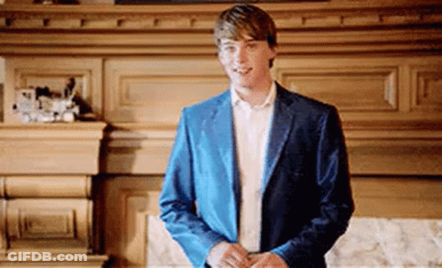A Young Man In A Blue Jacket And White Shirt Is Standing In Front Of A Fireplace . GIF