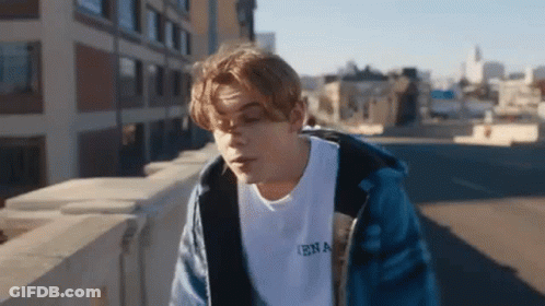 A Young Man In A Blue Jacket Is Standing On A Bridge . GIF