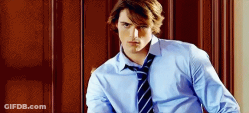 A Young Man In A Blue Shirt And Tie Is Standing In Front Of A Wooden Door . GIF
