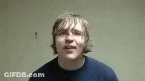 A Young Man In A Blue Shirt Is Making A Funny Face While Standing In Front Of A Wall . GIF