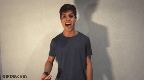 A Young Man In A Blue Shirt Is Standing In Front Of A White Wall And Making A Funny Face . GIF