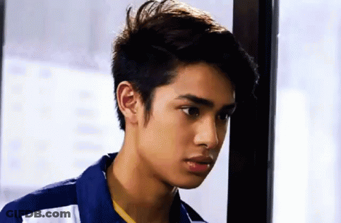 A Young Man In A Blue Shirt Is Standing Next To A Window And Looking At The Camera . GIF