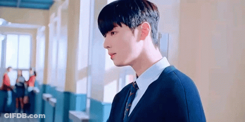 A Young Man In A Blue Sweater And Tie Is Standing In A Hallway . GIF