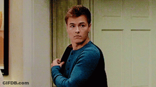A Young Man In A Blue Sweater Is Standing In Front Of A Door Holding His Jacket . GIF