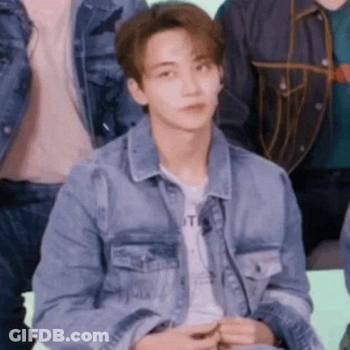 A Young Man In A Denim Jacket Is Sitting In A Group Of People . GIF