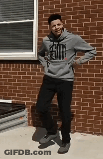 A Young Man In A Gray Hoodie Is Standing In Front Of A Brick Building . GIF