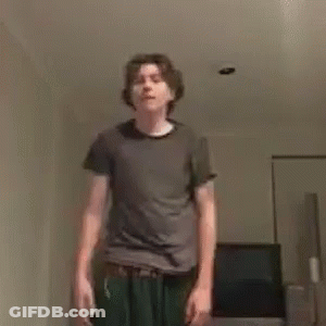 A Young Man In A Gray Shirt And Green Pants Is Standing In A Living Room . GIF