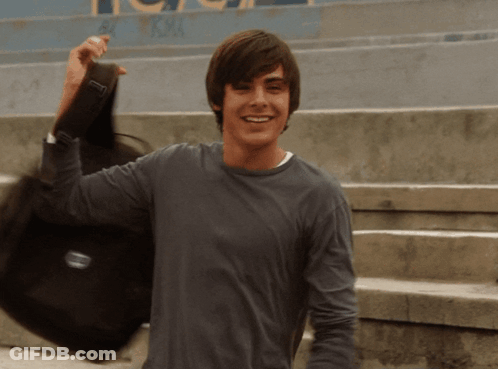 A Young Man In A Gray Shirt Is Holding A Black Bag With The Letter T On It GIF