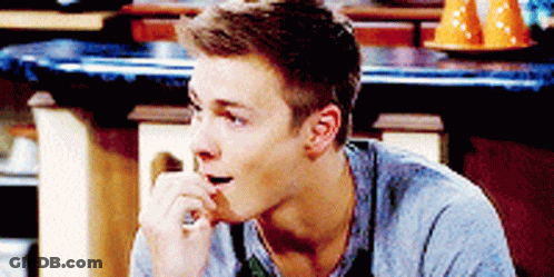A Young Man In A Gray Shirt Is Sitting At A Counter With His Hand On His Mouth GIF