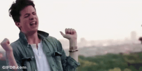 A Young Man In A Green Jacket And White Shirt Is Standing In Front Of A City With His Arms In The Air . GIF