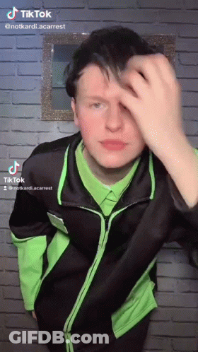 A Young Man In A Green Shirt And Black Jacket Is Standing In Front Of A Brick Wall . GIF