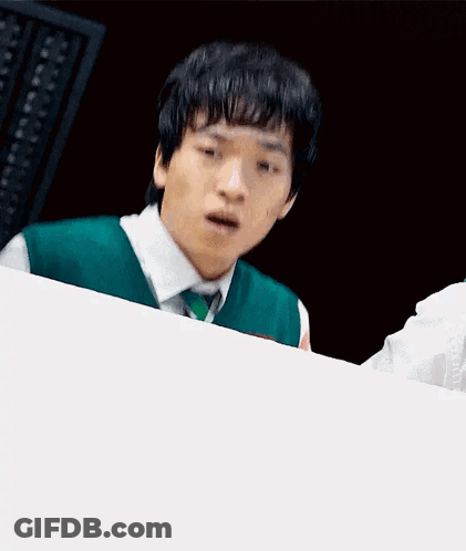 A Young Man In A Green Vest And Tie Is Sitting At A Table With His Mouth Open . GIF
