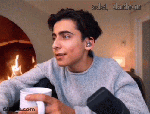 A Young Man In A Grey Sweater Is Holding A White Mug In Front Of A Fireplace GIF