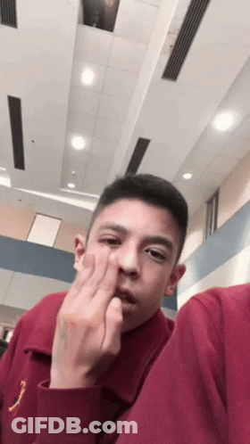 A Young Man In A Maroon Shirt Is Making A Funny Face While Sitting In A Room . GIF
