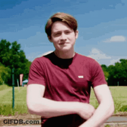 A Young Man In A Maroon Shirt Is Standing In A Field With His Arms Crossed . GIF