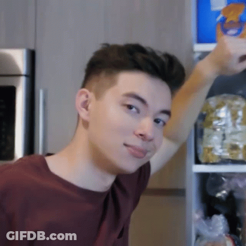 A Young Man In A Maroon Shirt Is Standing In Front Of A Refrigerator Reaching For Something GIF