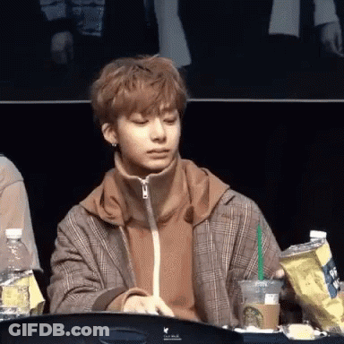 A Young Man In A Plaid Jacket Is Sitting At A Table Holding A Cup Of Coffee And A Bag Of Chips . GIF