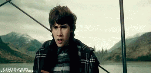 A Young Man In A Plaid Jacket Is Standing Next To A Body Of Water With Mountains In The Background . GIF