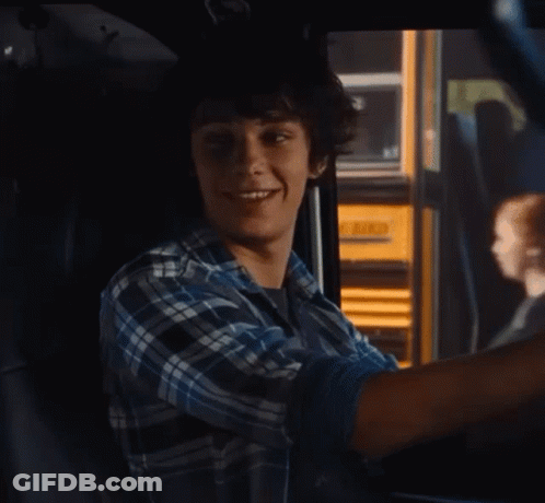 A Young Man In A Plaid Shirt Is Sitting In A Car With A Yellow School Bus In The Background GIF