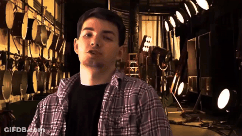 A Young Man In A Plaid Shirt Stands In Front Of A Wall Of Drums GIF