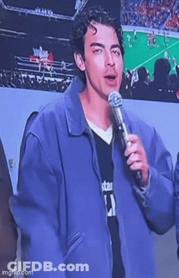 A Young Man In A Purple Jacket Is Holding A Microphone And Talking Into It . GIF