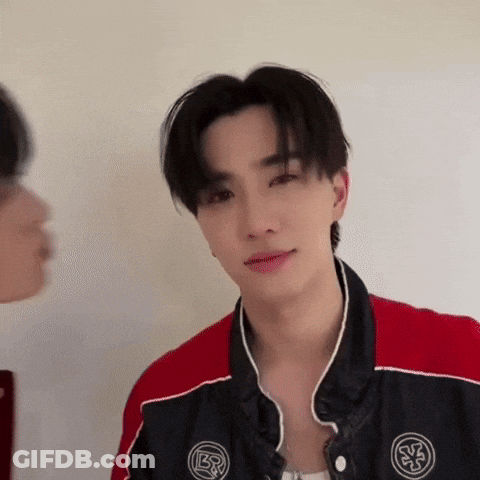 A Young Man In A Red And Black Jacket Is Looking At The Camera While Another Man Looks On . GIF