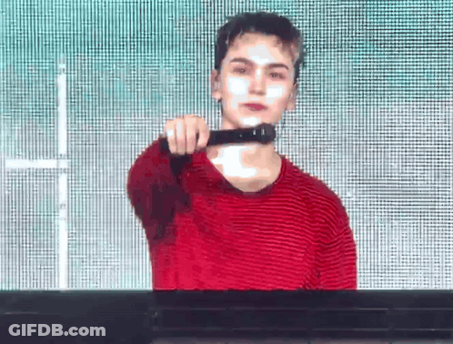A Young Man In A Red Striped Shirt Is Standing In Front Of A Screen And Holding A Microphone . GIF