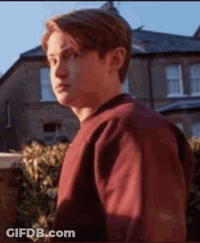 A Young Man In A Red Sweater Is Standing In Front Of A House And Looking At The Camera . GIF