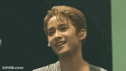 A Young Man In A Striped Shirt Is Smiling In Front Of A Microphone GIF