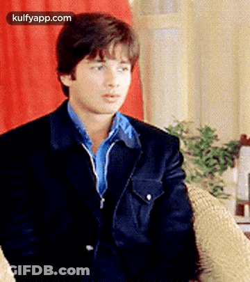 A Young Man In A Suit And Blue Shirt Is Sitting In A Chair . GIF