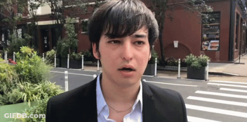 A Young Man In A Suit And Blue Shirt Is Standing In Front Of A Building . GIF