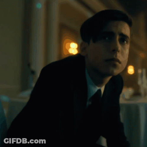 A Young Man In A Suit And Tie Is Sitting In A Dark Room . GIF