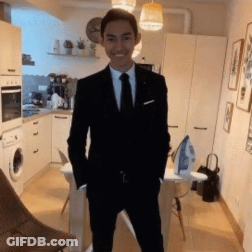 A Young Man In A Suit And Tie Is Standing In A Kitchen With His Hands In His Pockets . GIF