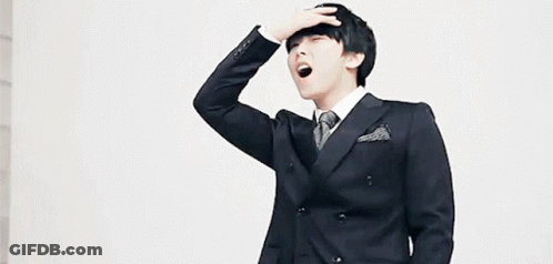 A Young Man In A Suit And Tie Is Yawning While Holding His Hand To His Forehead . GIF